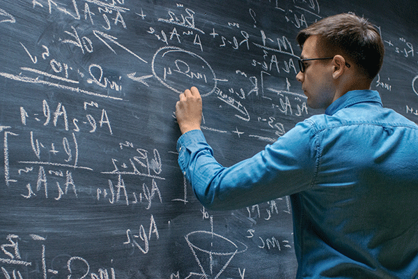 A man in a blue, long sleeved, collared shirt, who has short brown hair and wears glasses, writes equations on a chalk board that is full of them. 