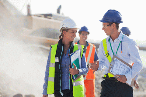 Two individuals are walking at a construction site. On the left is a white male with short brown hair. He holds a clipboard with a white paper document in his right hand. On the right is a white female with blond hair pulled back into a ponytail. The two individuals are looking at each other as they walk. Behind them is a white male wearing a blue hard hat and an orange full-body safety outfit with a blue hard hat. A bulldozer is in the background. 